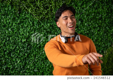 Asian man laughing and pointing by green wall, wearing orange sweatshirt and headphones, copy space Asian man laughing and pointing by green wall, wearing orange sweatshirt and headphones, copy space 141546512