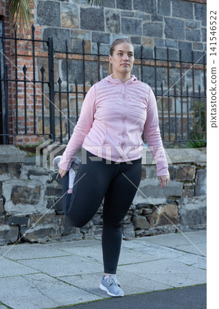 Woman in 20s stretching quads on sidewalk beside stone wall and iron fence wearing pink jacket Woman in 20s stretching quads on sidewalk beside stone wall and iron fence wearing pink jacket 141546522