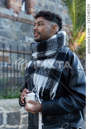Indian man standing on sidewalk by stone wall, wearing leather jacket and checked scarf holding cup 141546524