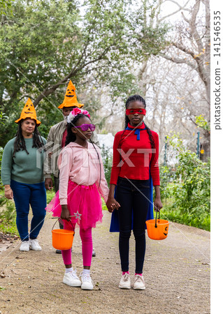 African American family walking on paved path, girls wearing cape, tutu, holding pumpkin buckets 141546535