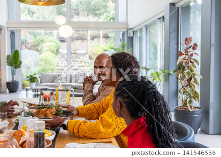 Diverse family with teenage daughter sharing roast chicken at table, mom in yellow sweater reaching Diverse family with teenage daughter sharing roast chicken at table, mom in yellow sweater reaching 141546536