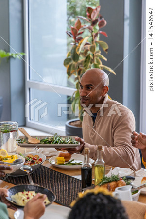 Indian man in beige pullover and friends serving green beans on platter with spatula at table Indian man in beige pullover and friends serving green beans on platter with spatula at table 141546542