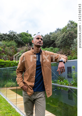 Asian man leaning on glass railing along boardwalk in garden beside pool, wearing smartwatch Asian man leaning on glass railing along boardwalk in garden beside pool, wearing smartwatch 141546543