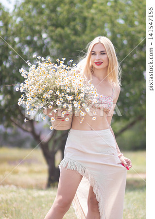 Smiling adult woman holds a pot of daisies and lightly lifts her skirt in a sunny meadow. Vertical summer portrait for lifestyle, fashion and floral themes. Smiling adult woman holds a pot of daisies and lightly lifts her skirt in a sunny meadow. Vertical summer portrait for lifestyle, fashion and floral themes. 141547156