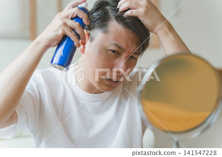 A man in his 40s is using hairspray to care for his hair while looking in the mirror. A man in his 40s is using hairspray to care for his hair while looking in the mirror. 141547228
