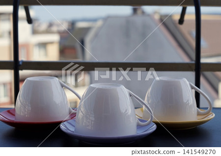 Three upside-down white cups sit on brightly colored saucers on a balcony table. The unusual arrangement symbolizes preparation, order, and quiet moments before serving drinks Three upside-down white cups sit on brightly colored saucers on a balcony table. The unusual arrangement symbolizes preparation, order, and quiet moments before serving drinks 141549270