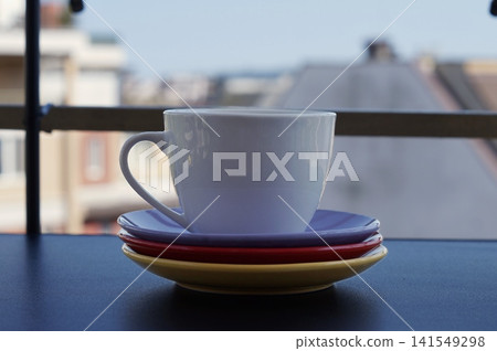 A white ceramic cup rests on a stack of purple, red, and yellow saucers near a balcony window. The colorful still life symbolizes simple morning rituals, comfort, and cheerful home hospitality A white ceramic cup rests on a stack of purple, red, and yellow saucers near a balcony window. The colorful still life symbolizes simple morning rituals, comfort, and cheerful home hospitality 141549298