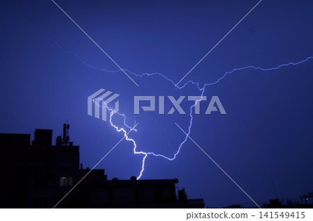 Brilliant forked lightning illuminates a deep blue night sky above black urban silhouettes. The image represents raw power, urgency, and a dramatic moment in nature Brilliant forked lightning illuminates a deep blue night sky above black urban silhouettes. The image represents raw power, urgency, and a dramatic moment in nature 141549415
