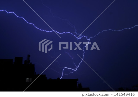 Bright branching lightning illuminates a dark blue night sky above silhouetted city buildings. Dramatic thunderstorm scene represents natural power, danger, and fleeting energy. Bright branching lightning illuminates a dark blue night sky above silhouetted city buildings. Dramatic thunderstorm scene represents natural power, danger, and fleeting energy. 141549416