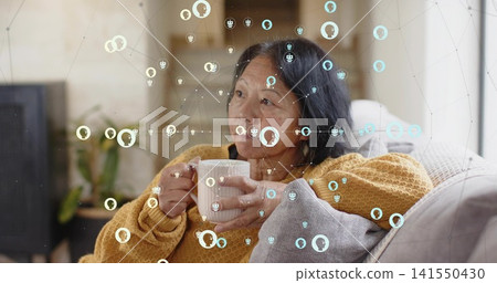 Sitting senior Chinese woman holding white mug on sofa at home in mustard sweater, net overlay Sitting senior Chinese woman holding white mug on sofa at home in mustard sweater, net overlay 141550430