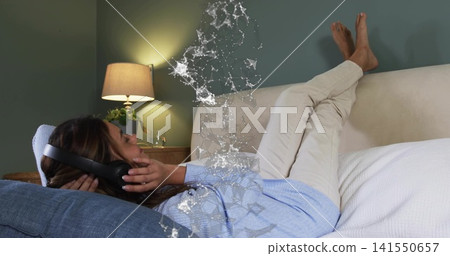 Lying woman in blue sweater and pants on white bed holding over-ear headphones with digital overlay Lying woman in blue sweater and pants on white bed holding over-ear headphones with digital overlay 141550657