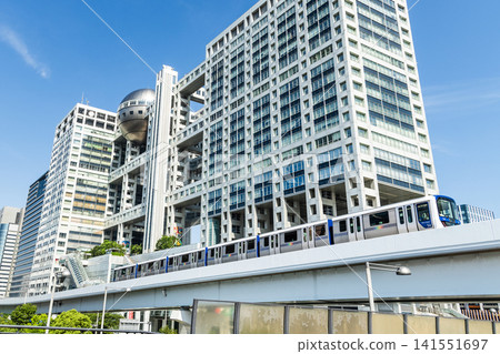 View of a New Transit Yurikamome line train running on the elevated track, driving past the Fuji Television building in Koto, Tokyo, Japan. It's an automated guideway transit service in Tokyo. View of a New Transit Yurikamome line train running on the elevated track, driving past the Fuji Television building in Koto, Tokyo, Japan. It's an automated guideway transit service in Tokyo. 141551697
