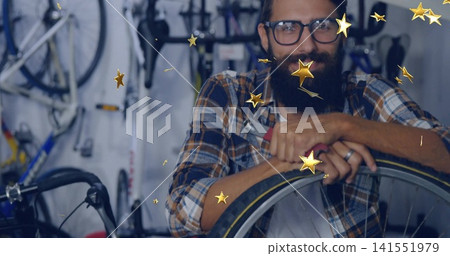 Leaning bearded mechanic in plaid shirt at bike shop, holding tire lever on wheel, gold overlays Leaning bearded mechanic in plaid shirt at bike shop, holding tire lever on wheel, gold overlays 141551979