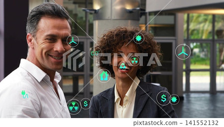 Standing two execs presenting energy visuals in atrium, with floating green icons and formal wear 141552132