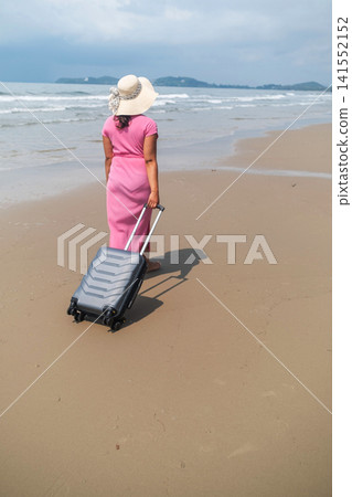 Young woman in pink dress and straw hat dragging a grey suitcase across wet reflective beach sand with sea waves in the background. Beach arrival concept Young woman in pink dress and straw hat dragging a grey suitcase across wet reflective beach sand with sea waves in the background. Beach arrival concept 141552152