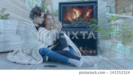 Cuddling two women in knit sweaters, jeans, under blanket in living room holding mug near fireplace 141552324