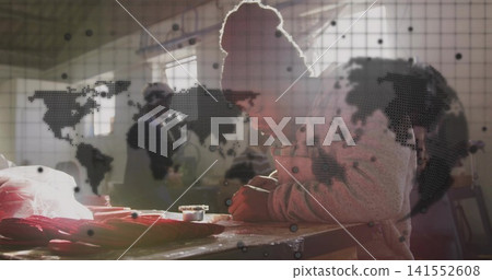 Leaning male wearing beanie inspecting red fabric at worktable by window, holding jar, map overlay Leaning male wearing beanie inspecting red fabric at worktable by window, holding jar, map overlay 141552608