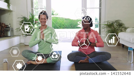 Meditating two women wearing activewear at home on black yoga mats, with water bottles and plants Meditating two women wearing activewear at home on black yoga mats, with water bottles and plants 141552648