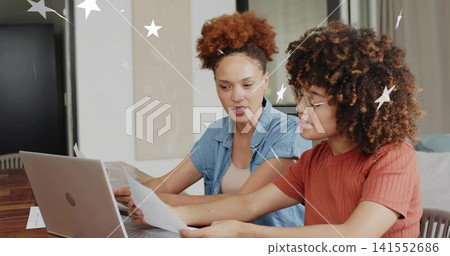 Reviewing papers, two women wearing denim shirt and rust top, glasses, using silver laptop at home 141552686