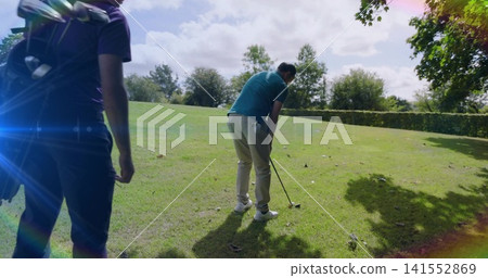 Preparing golfer in teal polo khakis bending to strike golf ball on fairway with golf bag 141552869