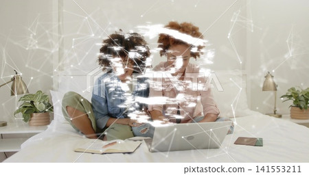 Watching two women in denim and pink shirts using silver laptop on bed, with notebook nearby 141553211