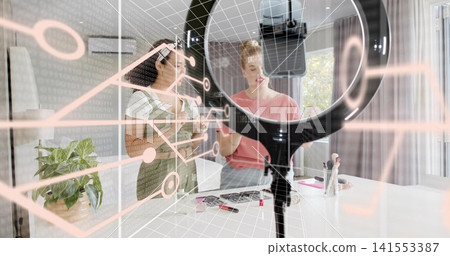 Demonstrating two women applying makeup at home workspace, with ring light, smartphone, AR overlay Demonstrating two women applying makeup at home workspace, with ring light, smartphone, AR overlay 141553387