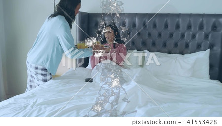 Leaning woman in blue handing orange juice to seated woman in pink in bed, molecular overlay Leaning woman in blue handing orange juice to seated woman in pink in bed, molecular overlay 141553428