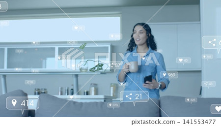Standing woman in denim shirt holding smartphone and mug in kitchen with floating icons, copy space Standing woman in denim shirt holding smartphone and mug in kitchen with floating icons, copy space 141553477