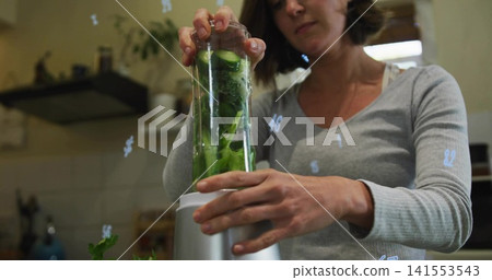 Fitting woman wearing grey top securing clear blender jar on counter with leafy greens and cucumber Fitting woman wearing grey top securing clear blender jar on counter with leafy greens and cucumber 141553543