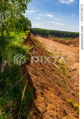 Steep sandy cliff edge overlooking a vast quarry pit with tall grass and lush trees under a clear blue summer sky, capturing a rugged and wild nature landscape scenery Steep sandy cliff edge overlooking a vast quarry pit with tall grass and lush trees under a clear blue summer sky, capturing a rugged and wild nature landscape scenery 141553671