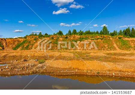 Exhausted sandy open-pit mine with eroded soil heaps, muddy puddle reflecting the bright blue sky and lush green forest edge on a warm summer day Exhausted sandy open-pit mine with eroded soil heaps, muddy puddle reflecting the bright blue sky and lush green forest edge on a warm summer day 141553687