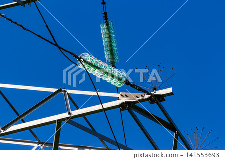 Close-up of glass high-voltage power line insulators on a steel transmission tower against a clear blue sky, showing industrial electrical equipment and metal construction details Close-up of glass high-voltage power line insulators on a steel transmission tower against a clear blue sky, showing industrial electrical equipment and metal construction details 141553693