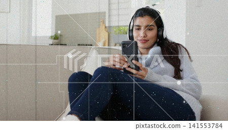Holding phone Indian woman wearing gray sweater and jeans scrolling phone on sofa, with headphones Holding phone Indian woman wearing gray sweater and jeans scrolling phone on sofa, with headphones 141553784