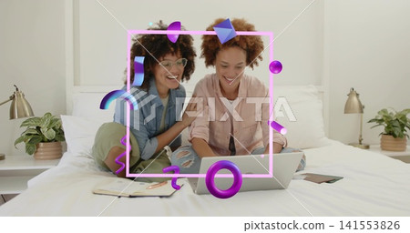 Leaning two women using laptop on bed at home, wearing denim and pink shirts, with overlay Leaning two women using laptop on bed at home, wearing denim and pink shirts, with overlay 141553826