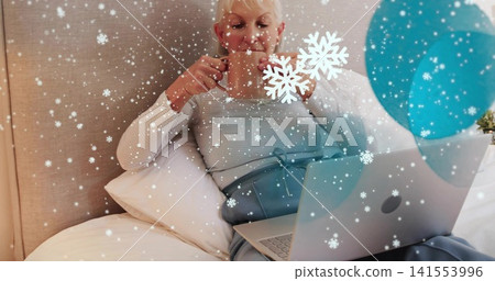 Reclining senior woman in striped top, sipping mug, typing on laptop in bedroom with turquoise disc Reclining senior woman in striped top, sipping mug, typing on laptop in bedroom with turquoise disc 141553996