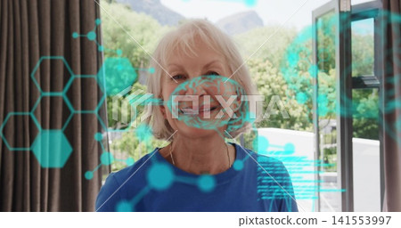 Standing senior woman in blue top and gold necklace at open glass door, teal holographic overlay Standing senior woman in blue top and gold necklace at open glass door, teal holographic overlay 141553997