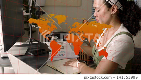 Writing woman in green pinafore taking notes at office, with laptop, monitor and orange map overlay 141553999