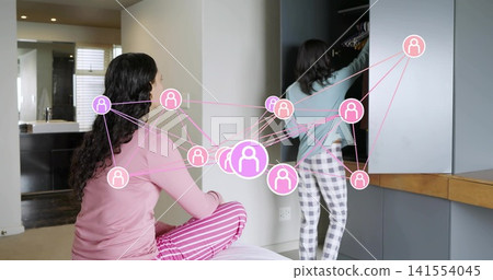 Sitting and reaching two women in pajamas organizing wardrobe in bedroom, with pink network overlay Sitting and reaching two women in pajamas organizing wardrobe in bedroom, with pink network overlay 141554045