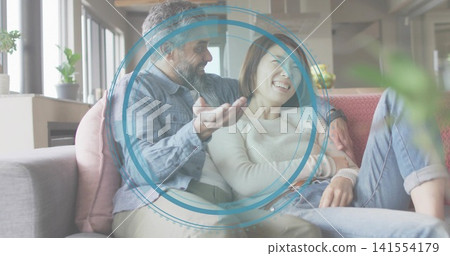 Sitting denim-shirt and cream-sweater couple cuddling on sofa at home, blue overlay, pink pillow Sitting denim-shirt and cream-sweater couple cuddling on sofa at home, blue overlay, pink pillow 141554179