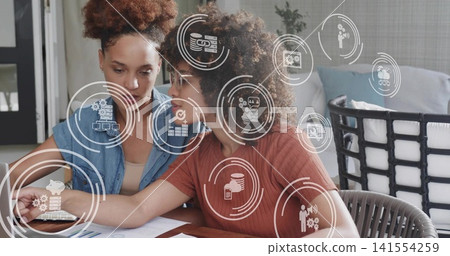 Reviewing documents, two women in rust, blue tops discussing charts at home with finance icons Reviewing documents, two women in rust, blue tops discussing charts at home with finance icons 141554259