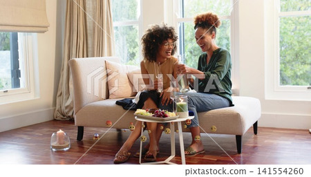 Pouring woman in green blouse and friend clinking glasses, sitting on beige loveseat with fruit Pouring woman in green blouse and friend clinking glasses, sitting on beige loveseat with fruit 141554260