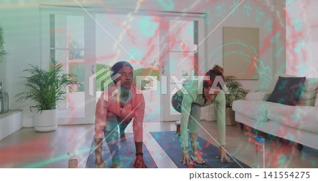 Practicing two women holding cat-cow on mats in living room, wearing pink and green tops Practicing two women holding cat-cow on mats in living room, wearing pink and green tops 141554275