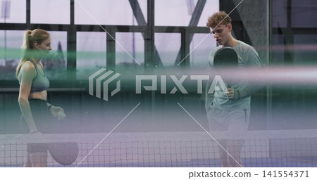 Playing players at net on court, woman in sports bra, man in long-sleeve top holding paddles Playing players at net on court, woman in sports bra, man in long-sleeve top holding paddles 141554371