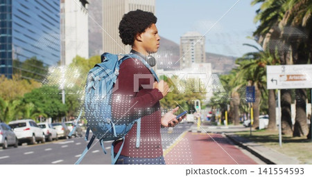 Standing man wearing maroon sweater checking phone on city road with blue backpack, headphones Standing man wearing maroon sweater checking phone on city road with blue backpack, headphones 141554593
