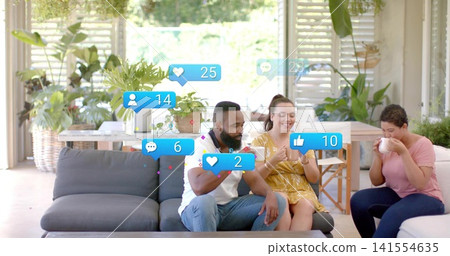 Smiling woman in yellow dress showing phone, friends sipping mugs on gray sofa with floating alerts Smiling woman in yellow dress showing phone, friends sipping mugs on gray sofa with floating alerts 141554635