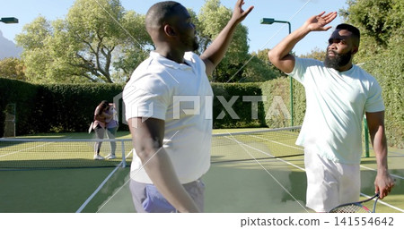 High-fiving two men in white polo and mint T-shirt on tennis court, with racket and smartphone High-fiving two men in white polo and mint T-shirt on tennis court, with racket and smartphone 141554642