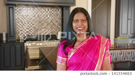 Smiling Indian woman standing at kitchen island, wearing saree and gold necklace, gas range behind 141554663