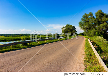 Asphalt country road winding through sunny summer fields with lush green trees and a quiet river under a vast clear blue sky on a bright day 141555179