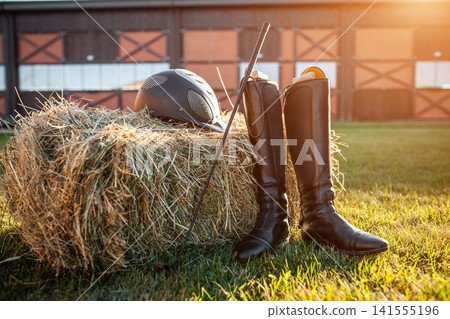 equestrian horse riding equipment with helmet, leather boots and stick on grass and hay bale, barn yard and rancho on background, sunny shot equestrian horse riding equipment with helmet, leather boots and stick on grass and hay bale, barn yard and rancho on background, sunny shot 141555196