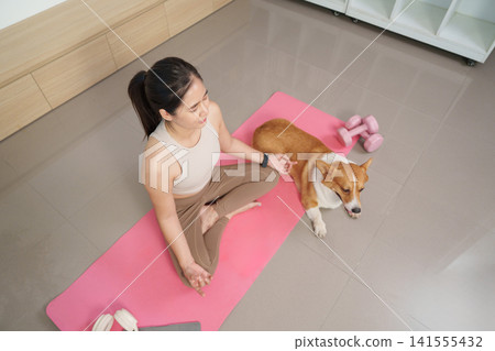 Asian woman practicing yoga and meditating on pink mat with corgi dog at home 141555432